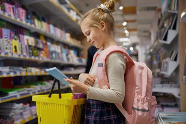 Küçük kız öğrenci süpermarket kırtasiyesinde alışveriş yapıyor. Okulda okumak için kitap alan sevimli küçük kız modern dükkanda alışverişin keyfini çıkarıyor.