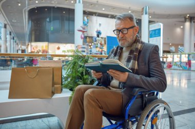 Sandalyesini rahat bir şekilde bankta oturan ve günün tadını çıkarırken ilginç bir kitap okuyan bir adam.