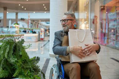 Tekerlekli sandalyede oturmuş alışveriş merkezinin içinde alışveriş çantası taşıyan bir adam.