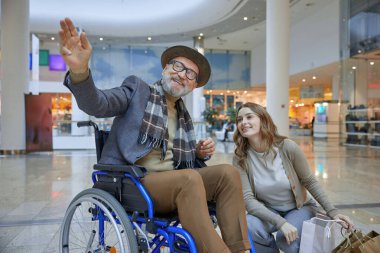Tekerlekli sandalye kullanan bir adam, elinde birkaç poşet tutan bir kadının yanında rahatça oturuyor.
