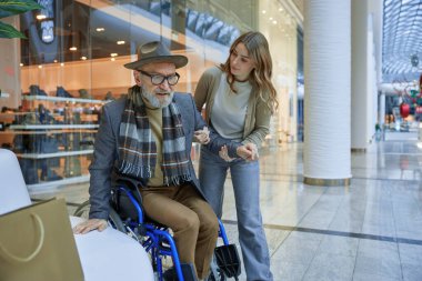 Tekerlekli sandalyede engelli bir adam, alışveriş merkezinden geçen bir bayandan yardım alıyor.