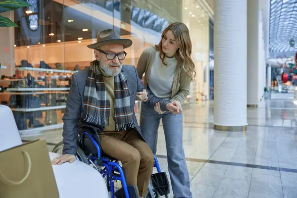 Tekerlekli sandalyede engelli bir adam, alışveriş merkezinden geçen bir bayandan yardım alıyor.