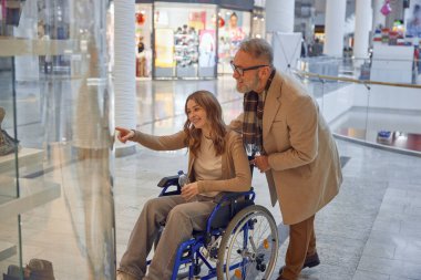 Alışveriş merkezinde neşeli bir sahne. Tekerlekli sandalyedeki bir kız, yaşlı bir adamın ona yardım ettiği vitrindeki sergiye hayran kalıyor.