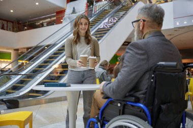 Bir alışveriş merkezinde engelli bir adama tekerlekli sandalyede yardım eden ve elinde kahve tutan genç bir bayan.