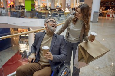 Zevkli bir alışveriş gününün tadını çıkarırken tekerlekli sandalyedeki bir adamı arkadaşının yanında gösteren neşeli bir sahne.