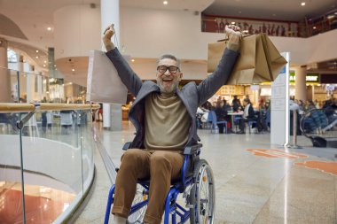 Tekerlekli sandalyedeki neşeli bir adam alışveriş merkezinden alışveriş yapıyor, elinde bir sürü alışveriş çantası taşıyor.