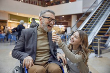 Tekerlekli sandalyedeki adam büyük bir alışveriş merkezinde yardım ediyor, dondurmanın tadını çıkarıyor.