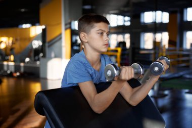Beden eğitiminde dambıllarla bisepsi pompalayan genç bir çocuk. Genç sporcu ağırlıklarla spor yapıyor. Spor ve vücut bakımında çocukların motivasyonu