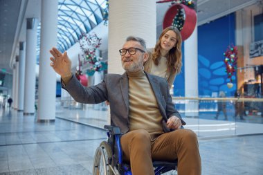 Kadın, tekerlekli sandalyedeki bir adamı yumuşak ışıkla dolu uzun bir koridorda nazikçe iter.
