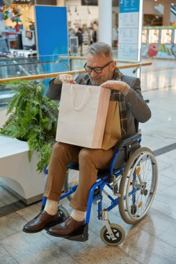 Tekerlekli sandalyede oturmuş alışveriş merkezinin içinde alışveriş çantası taşıyan bir adam.