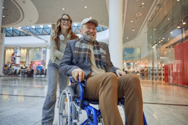 Kalabalık bir alışveriş merkezinde, merhametli bir torun tekerlekli sandalye kullanan dedesine yardım ediyor.