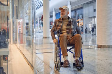 Tekerlekli sandalyede oturan yaşlı bir adam düşünceli bir şekilde modern bir alışveriş merkezinin modaya uygun atmosferinin tadını çıkarıyor.