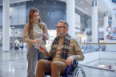 Alışveriş merkezinin hareketli ortamında, merhametli bir kadın, tekerlekli sandalye kullanan yaşlı bir adama yardım ediyor.