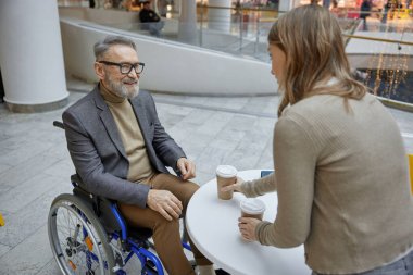 Alışveriş merkezinde engelli tekerlekli sandalyedeki olgun bir adama yardım eden, elinde kahve tutan genç bir bayan.