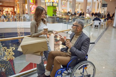 Bir kadın ve tekerlekli sandalyedeki yaşlı bir adam kalabalık bir alışveriş merkezinde kahve içerken bir an paylaşıyorlar.