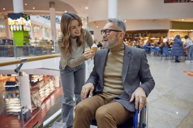 Tekerlekli sandalyedeki adam ve bayan asistanı büyük bir alışveriş merkezinde dondurma yiyor.
