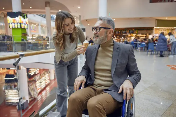 Tekerlekli sandalyedeki adam ve bayan asistanı büyük bir alışveriş merkezinde dondurma yiyor.
