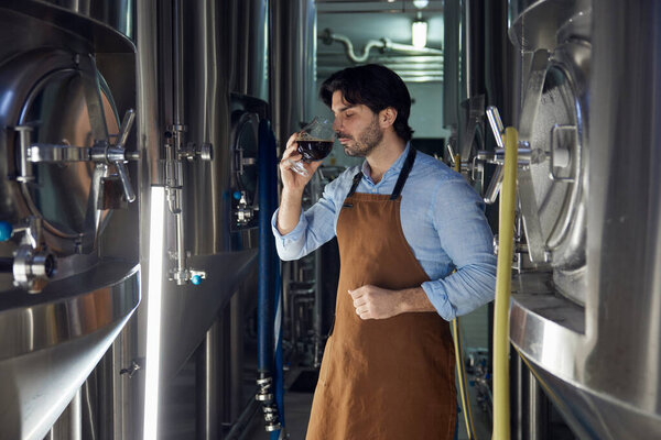 Человек в традиционном фартуке держит высокий, морозный стакан, наполненный освежающим пивом, возможно, из местной пивоварни или винодельни