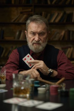 Yetişkin bir adam elinde puroyla eski bir kütüphanede kağıt oynuyor.