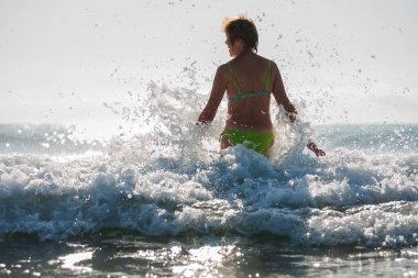 Sıska tenli, mayo giymiş bir bayan, güneşli bir havada dalgaların arasından denize giriyor. 