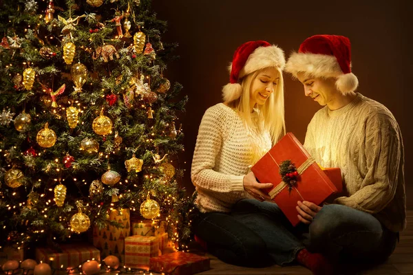 Christmas Couple opening Xmas Gift Box. Happy Couple in Santa Hat with Red Present next Christmas tree in Dark Room Interior. Winter Holiday Celebration at Home
