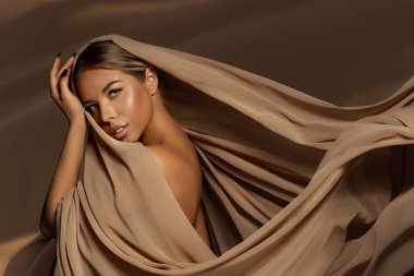 Doğal makyajlı ve bronzlaşmış tenli güzel kız yüzü. Kahverengi ipek kumaşa sarılmış güzel kadın rüzgarda sallanıyor. Baş Atkısı takan Moda Moda Portresi