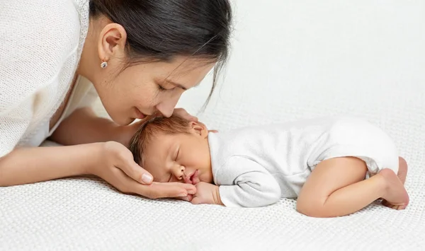 母亲亲吻睡在白色毛毯上的婴儿 笑着妈妈手牵着新生儿的头 婴儿侧视图躺在胃上 新生儿保健 母性快乐 — 图库照片