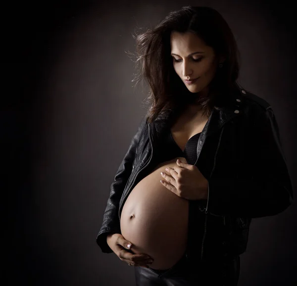 バイカーレザージャケットのスタイリッシュな妊婦は腹部を抱いています 妊娠中の腹側暗い背景にビュー マタニティファッションスタイルとヘルスケア 美しいですお母さん見腹以上グレー — ストック写真