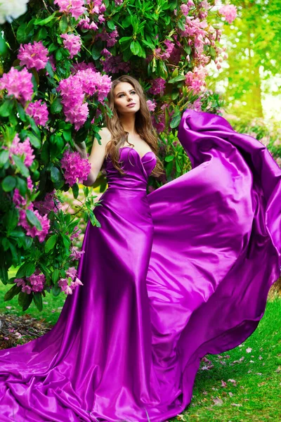 花の庭で長い紫のドレスで美しい夢の女性 春の公園で花の香りを楽しむファンタジーガール アウトドアとサテンピンクのガウンのファッションモデル — ストック写真