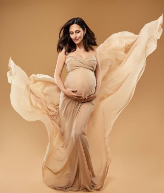 Bej Fantezi Elbiseli Güzel Hamile Kadın Rüzgâr Gibi Kanat çırpıp Göbeğe Bakıyor. Kahverengi Arkaplan 'ın üzerinde gecelikli güzel anne. Hamilelik kıyafetleri. Ana Sağlık Hizmetleri