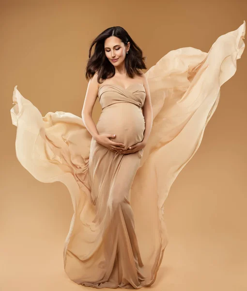 美しいベージュファンタジードレス風になびく妊娠中の女性はベリーを見て 茶色い背景の上のシフォンガウンの美しい母 妊娠中のファッション服 母子保健 — ストック写真