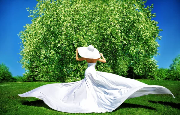 ファッション女性屋外で長い白いドレスリアビュー 白い夏の帽子の美しい女の子太陽を楽しむバックビュー 春の庭で花の木とエレガントな女性 — ストック写真