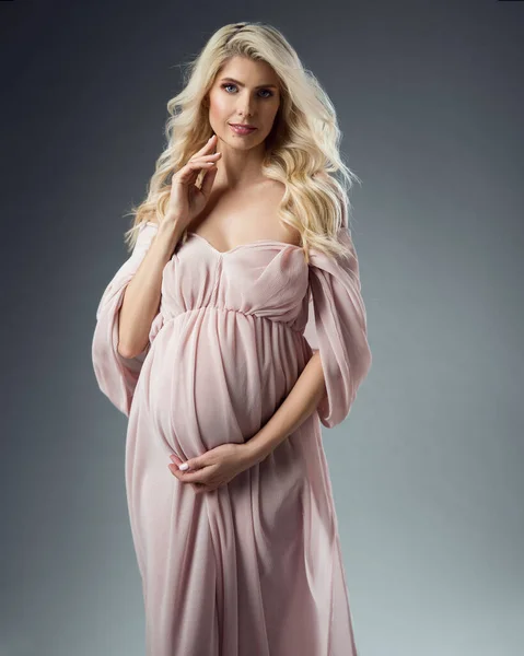 ピンクのシフォンの美しい妊婦の肖像は灰色の背景の上にドレスアップ シルクガウンにカーリーロングヘアで幸せな母 妊娠中のファッション — ストック写真