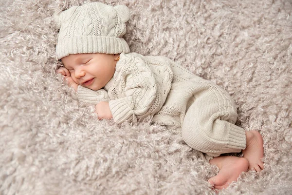 Newborn Baby smiling in Sleep on fluffy Blanket. One Month Child in knitted Hat Woolen Suit. Happy Infant Boy sleeping on Fur Bed