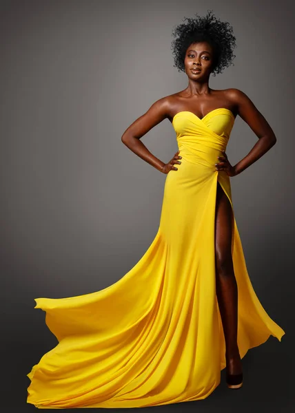 Fashion Dark Skin Woman in Yellow Silk Dress flying over Gray. African Model with Afro Curly Hair Style in Long Evening Gown. Stylish Lady Portrait