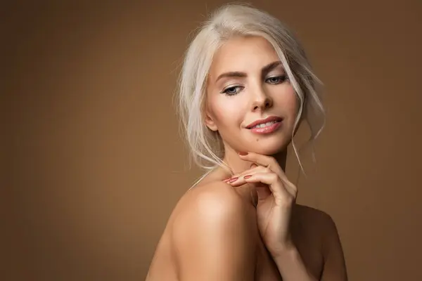 Beauty Model with nature Make up and blond Hair over beige. Beautiful smiling Woman with clean Skin touching Chin. Body Care and Face Lifting Treatment