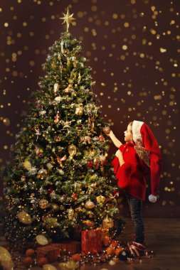 Işıklı Noel Ağacı. Noel Şapkalı Küçük Çocuk Noel Ağacı 'nı süsleyerek süslüyor. Parlak çelenklerle dolu samimi bir arka plan.