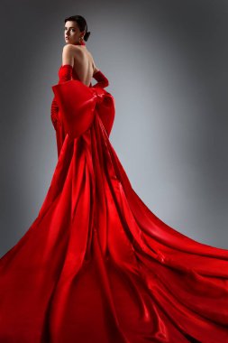Kırmızı İpek Sırtsız Elbiseli Moda Kadını. Gri Arkaplan Üzerinde Açık Sırt Maxi Elbisesi Giyen Zarif Kadın. Uzun İpek Tren Kumaşlı Güzel Gelinlik Modeli