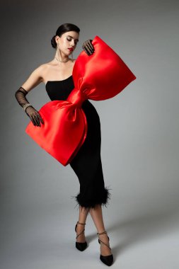Siyah elbiseli zarif bayan, Büyük Kırmızı Fiyonk 'u tutuyor. Gri Arkaplan üzerinde kurdele kemeri olan Kokteyl Elbiseli Moda Kızı. Yetenekli Güzel Kadın. Kadın Balosu Partisi Şık Kıyafetler