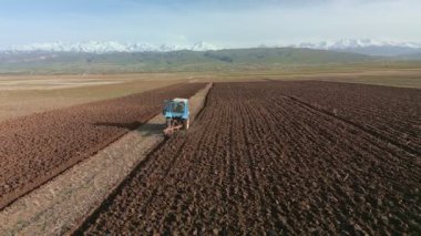 Blue Tractors sowing on agricultural field in spring, readying the field for planting. Powerful tractor turns the soil against the backdrop of high mountains. Issyk-kul Region valley in Kyrgyzstan, Tien Shan mountains.