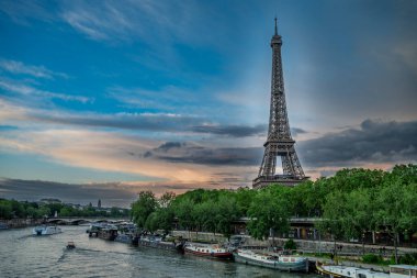 Paris 'teki Seine Nehri' nin üzerinde yükselen Eyfel Kulesi 'nin manzarası. Nehir kıyısı boyunca yanaşmış tekneler ve tepemizde parlak bir gökyüzü var..