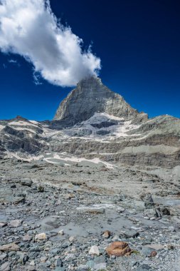 Kısmen bulutlarla kaplı ikonik Matterhorn dağı, buzul kalıntılarıyla kayalık bir arazinin üzerinde yükselir..