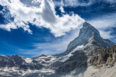 Matterhorn 'un çarpıcı manzarası mavi gökyüzünün altında dağınık bulutlarla görkemli zirvesini ve engebeli arazisini sergiliyor..