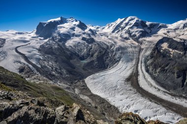 Panoramik bir manzara, açık mavi gökyüzünün altında karmaşık buz oluşumları ve çevresindeki zirveleriyle geniş Matterhorn buzullarını sergiliyor..