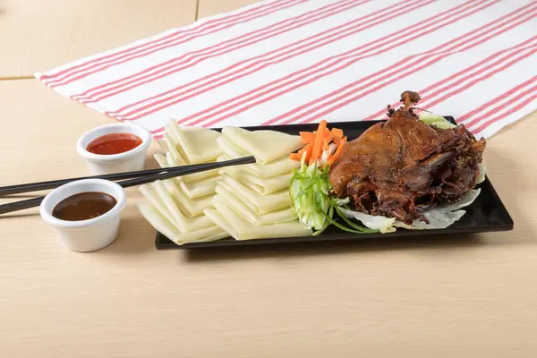 stock image A traditional Asian dish, crispy duck is served with rice sheets, vegetables and dipping sauces on a wooden table, with a stripe towel in the background