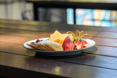 Beyaz bir tabakta servis edilen çeşitli peynirler, krakerler, meyveler, turşular ve kurutulmuş meyveler içeren gurme peynir tabağı.