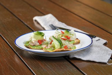 Üstünde Queensland kralbalığı, avokado sosu, salatalık kurdelesi, acı biber dilimleri ve dereotlu garnitür olan gevrek bir ekmek.