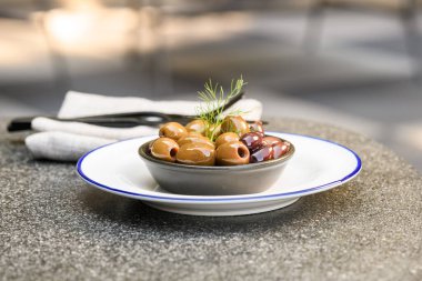 Taze dereotuyla süslenmiş çeşitli sıcak zeytinler, mavi kenarlı beyaz bir tabakta siyah bir kasede servis ediliyor.