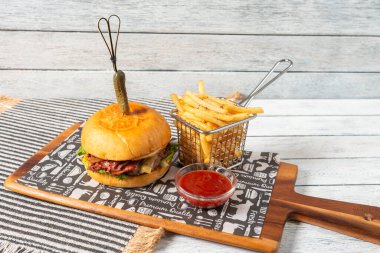Juicy hamburger topped with fresh ingredients sits on a wooden platter. Accompanied by crispy fries in a metal basket and tangy sauce, creating a hearty meal experience.