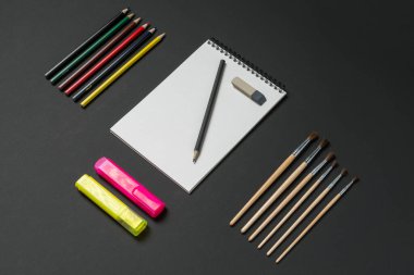 Karanlık bir yüzeyde, renkli kalemler, fosforlu kalemler, boya fırçaları, silgi ve boş bir eskiz kitabı, yaratıcı bir çalışma alanında sanatsal çabalar için ilham kaynağı olur..